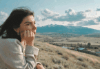 Uma mulher jovem está sentada em um ambiente aberto e de natureza com montanhas. Ela contempla a paisagem.