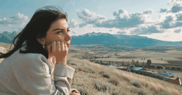 Uma mulher jovem está sentada em um ambiente aberto e de natureza com montanhas. Ela contempla a paisagem.