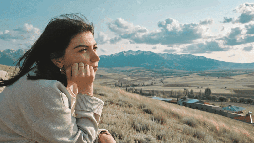 Uma mulher jovem está sentada em um ambiente aberto e de natureza com montanhas. Ela contempla a paisagem.