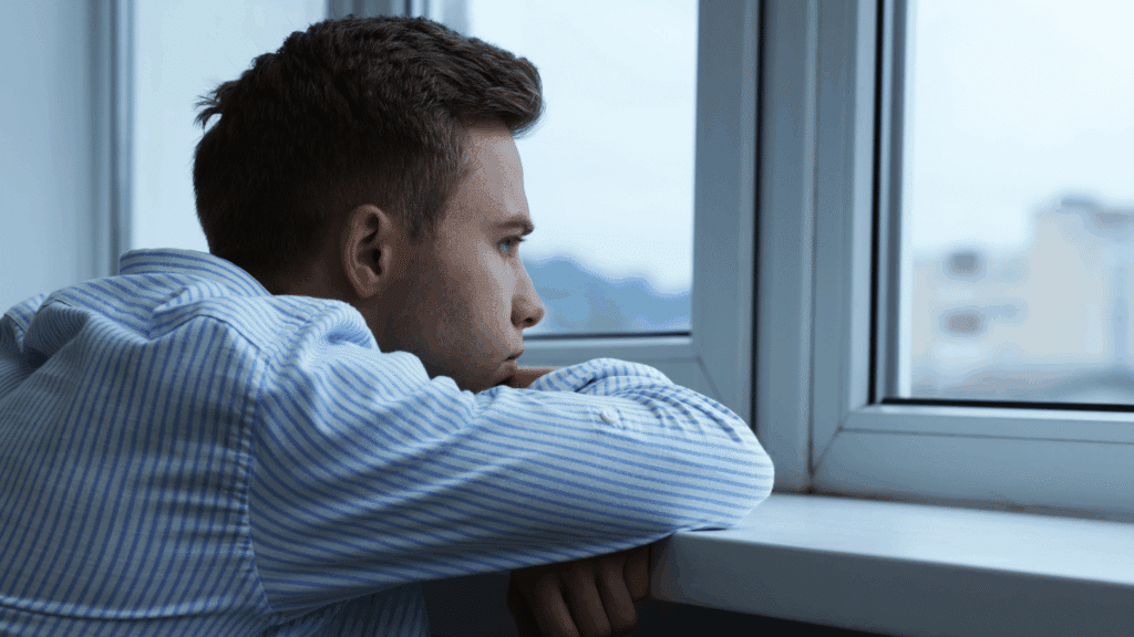Um homem jovem está atrás de uma janela em um ambiente fechado, dentro de uma casa. Ele observa pela janela e está encostado nela.