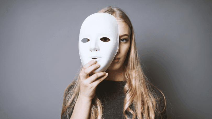 Uma mulher jovem está escondendo seu rosto atrás de uma máscara branca.