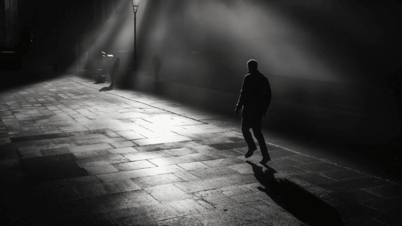 Um homem está caminhando por uma rua molhada à noite. O ambiente está escuro e há um poste refletindo um pouco de luz no local.