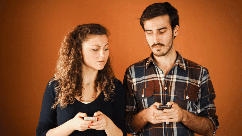 Mulher e homem lado a lado segurando celulares, olhando para baixo com expressões sérias ou desconfiadas, diante de um fundo laranja.