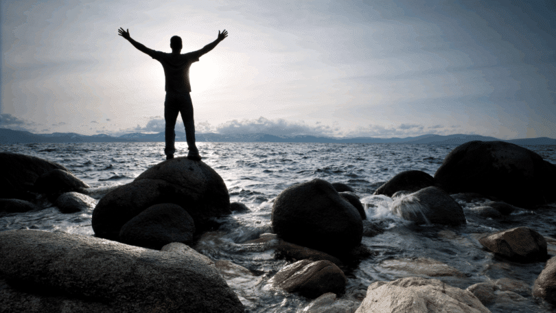Um homem está em pé em cima de uma pedra e de frente para o mar. Ele levanta os braços e observa o sol e o ambiente.