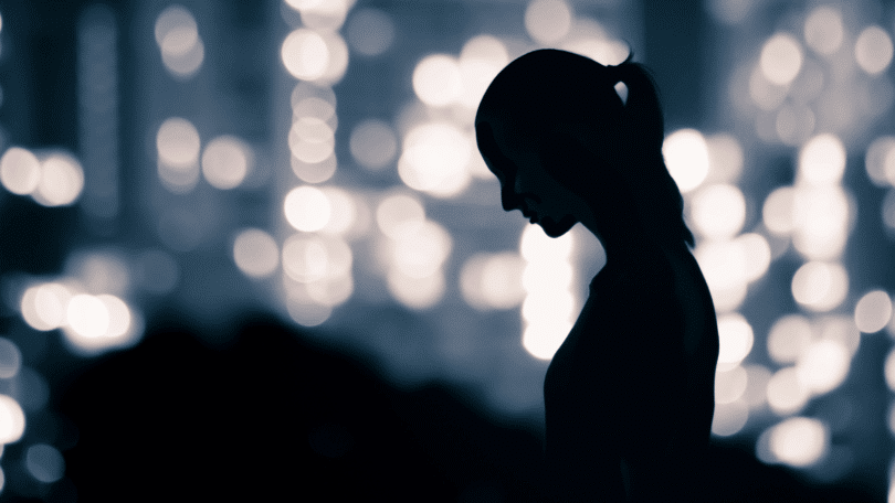 A silhueta de uma mulher cabisbaixa está em destaque na imagem. Ao fundo, há luzes da cidade.