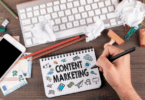 Mão segurando uma caneta escreve em um caderno com a frase ‘Content Marketing’, cercada por ícones de comunicação e marketing, sobre uma mesa com teclado, celular, papéis amassados, lápis e materiais de escritório.
