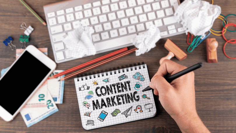 Mão segurando uma caneta escreve em um caderno com a frase ‘Content Marketing’, cercada por ícones de comunicação e marketing, sobre uma mesa com teclado, celular, papéis amassados, lápis e materiais de escritório.