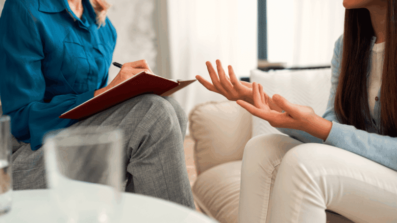 Uma adolescente está em uma sessão de terapia com a sua psicóloga. A profissional faz anotações no seu caderno, enquanto a paciente fala.