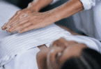 As mãos de um profissional fazendo reiki em uma cliente estão em destaque na imagem.