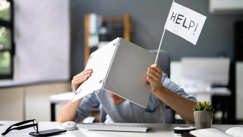 Imagem de um homem sentando. Ele está em seu ambiente de trabalho segurando um notebook e uma bandeira com a palavra Help. Ele tem o sintoma do Transtorno da Sobrecarga Sensorial.