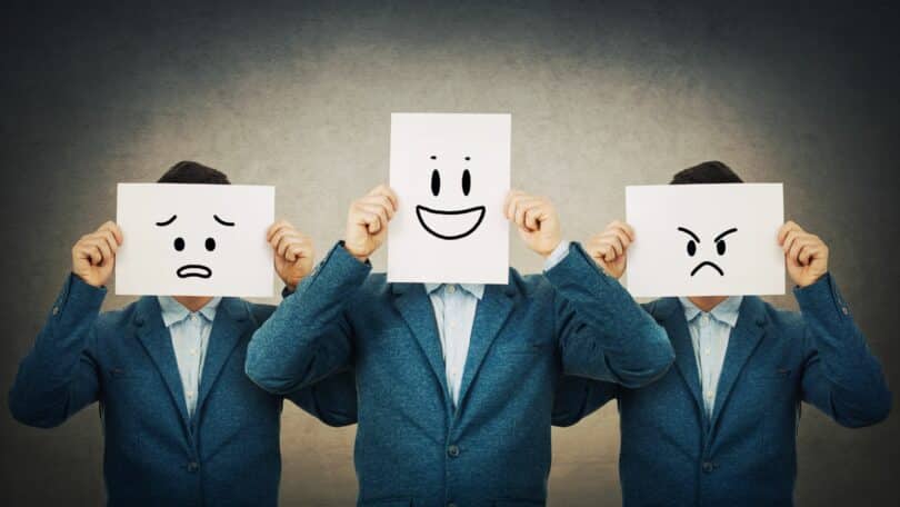 Imagem de três homens usando terno e camisa azul. Cada um segura um cartaz com um emoji diferente, trazendo o simbolismo de cada emoção sentida por eles.