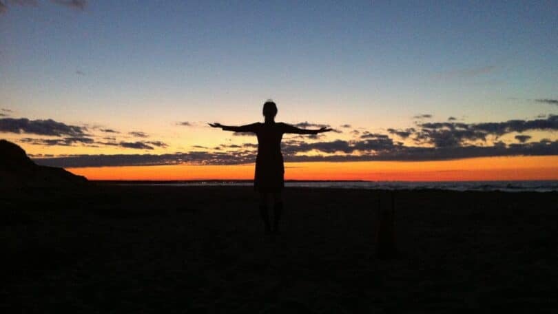 Imagem da silhueta de uma mulher de braços abertos para o pôr do Sol em frente ao mar.