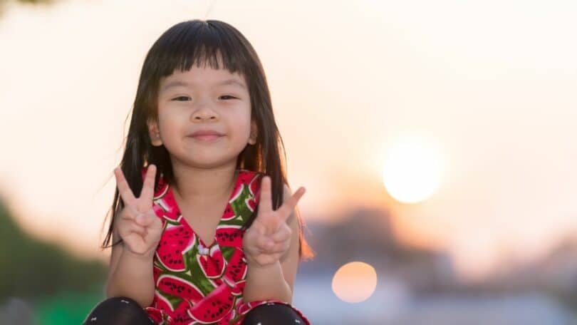 Imagem de uma criança mestiça sorrindo e com os dedos das mãos em sinal de paz. Ao fundo, um lindo pôr do Sol.