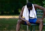 Imagem de um atleta negro, corredor, sentado em um banco de madeira pintado na cor verde. Ele está se sentindo desolado, com uma toalha branca no pescoço. A imagem simboliza o conceito de um vazio depois da conquista.