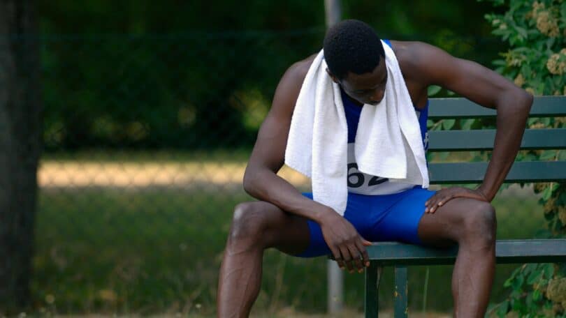 Imagem de um atleta negro, corredor, sentado em um banco de madeira pintado na cor verde. Ele está se sentindo desolado, com uma toalha branca no pescoço. A imagem simboliza o conceito de um vazio depois da conquista.