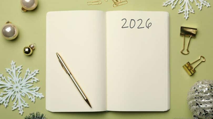 Imagem de um caderno com a indicação do ano 2026 no cabeçalho e uma caneta dourada sobre ele. Ao lado, bolas de Natal e outros enfeites nas cores dourada e prata.