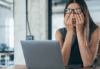 Uma mulher de negócios está sentada na frente de seu notebook. Ela coloca as duas mãos no rosto, indicando estar cansada.