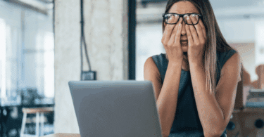 Uma mulher de negócios está sentada na frente de seu notebook. Ela coloca as duas mãos no rosto, indicando estar cansada.