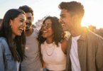 Um grupo de quatro pessoas, composto por duas mulheres e dois homens, estão sorrindo e olhando entre si. Eles estão em um ambiente ao ar livre.