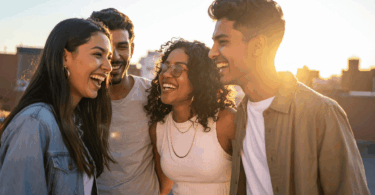 Um grupo de quatro pessoas, composto por duas mulheres e dois homens, estão sorrindo e olhando entre si. Eles estão em um ambiente ao ar livre.