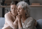 Uma mulher adulta e uma mulher idosa, que são filha e mãe, se confortam. Uma está segurando a mão da outra e elas estão de olhos fechados, indicando estarem chateadas.