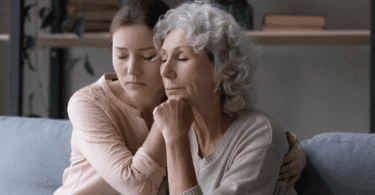 Uma mulher adulta e uma mulher idosa, que são filha e mãe, se confortam. Uma está segurando a mão da outra e elas estão de olhos fechados, indicando estarem chateadas.