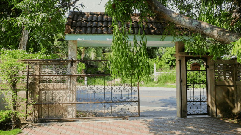 Um portão de uma casa está sendo aberto. Ao redor dela, há várias árvores e plantas.