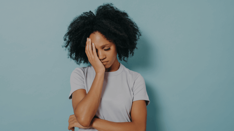 Mulher com a mão no rosto e expressão de tristeza ou preocupação, em fundo azul claro.