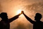 Imagem da silhueta de dois homens, de mãos dadas e levantadas e ao fundo um lindo pôr do Sol.