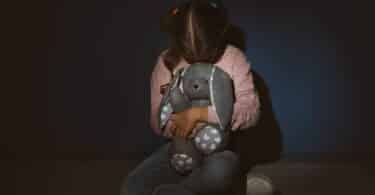 Imagem de uma menina assustada segurando um brinquedo, encostada perto de uma parede azul. A foto traz o conceito de Violência contra as crianças.