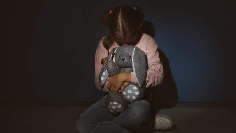 Imagem de uma menina assustada segurando um brinquedo, encostada perto de uma parede azul. A foto traz o conceito de Violência contra as crianças.