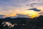 Há um ambiente de natureza na foto. O céu está amarelado do pôr do sol. Há um rio e diversas árvores e plantas ao redor. Há algumas pessoas sentadas em um morro.