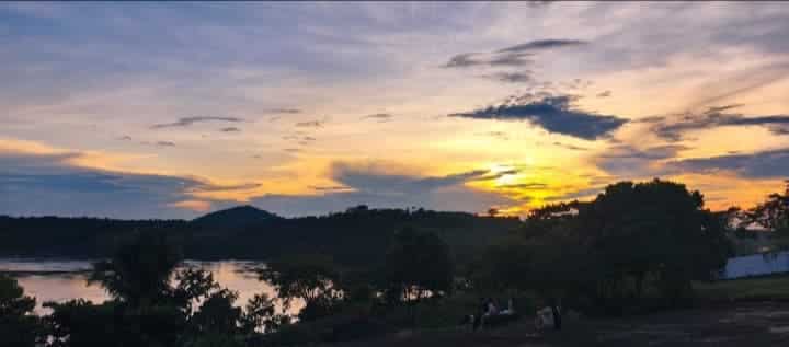 Há um ambiente de natureza na foto. O céu está amarelado do pôr do sol. Há um rio e diversas árvores e plantas ao redor. Há algumas pessoas sentadas em um morro.