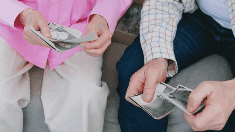Duas pessoas sentadas observando fotografias antigas em preto e branco, segurando as imagens com as mãos.