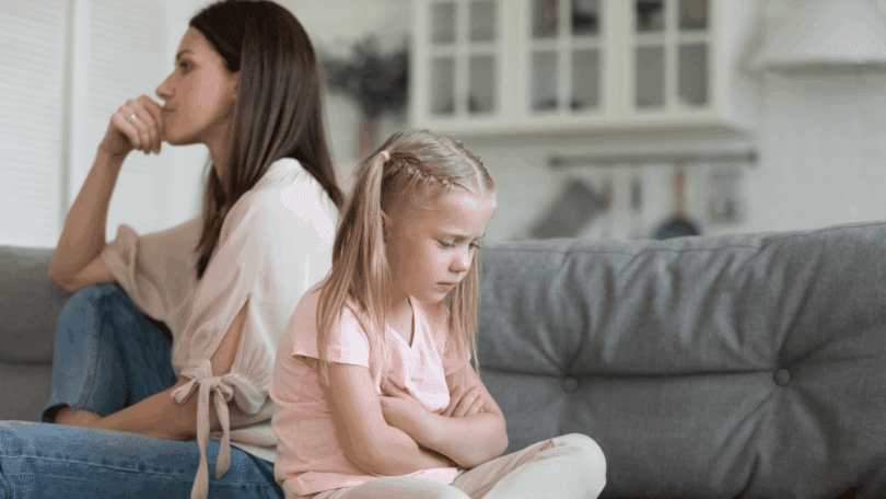 Uma mãe e uma filha criança estão de costas uma para a outra e sentadas em um sofá. A menina está chateada e a mãe está ignorando-a.