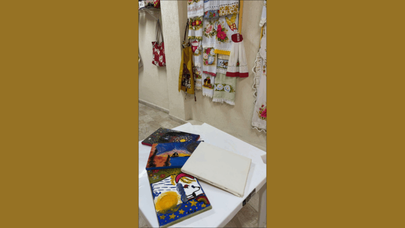Mesa branca com telas de pintura coloridas apoiadas, em um ambiente interno com panos e artesanato pendurados na parede ao fundo.