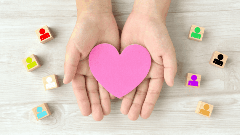 Mãos em concha seguram um coração rosa, cercadas por blocos com ícones de pessoas coloridas sobre uma superfície de madeira clara.