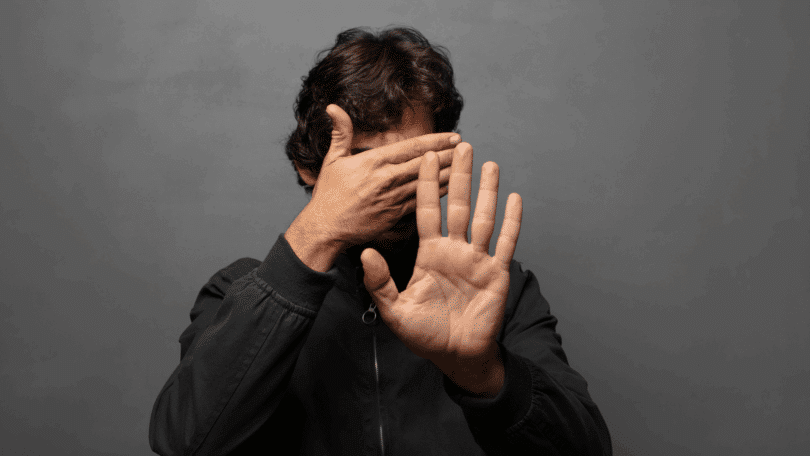 Homem cobre o rosto com uma mão enquanto estende a outra em gesto de proteção ou recusa, sobre fundo cinza.