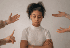 Mulher jovem de braços cruzados e olhar baixo, enquanto mãos ao redor apontam e gesticulam em sua direção.
