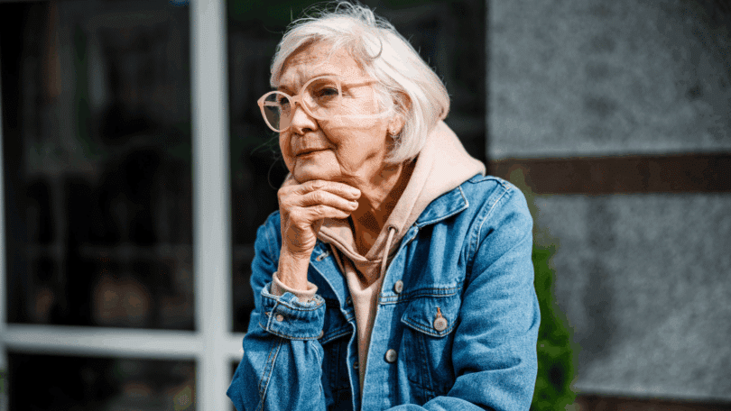 Uma mulher idosa está pensativa em um ambiente ao ar livre.