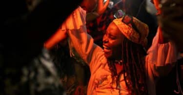 Imagem de uma mulher angolana, feliz e dançando ao som de uma música ao vivo, em uma área com várias pessoas.