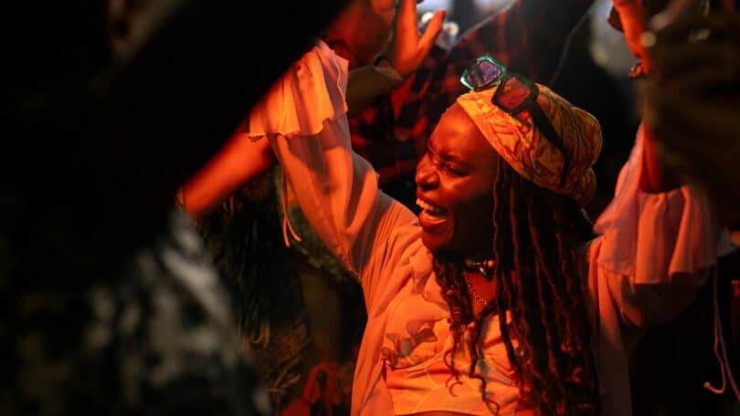 Imagem de uma mulher angolana, feliz e dançando ao som de uma música ao vivo, em uma área com várias pessoas.