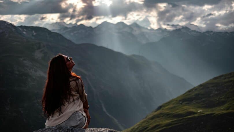 Imagem de uma mulher de cabelos longos e óculos de sol, sentada olhando para uma vista tranquila para a montanha nos Alpes Suíços.