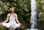 Imagem de uma mulher usando roupas brancas e uma rosa amarela no cabelo. Ela está fazendo a sua meditação e o seu mantra, sobre uma pedra, ao lado de uma pequena cachoeira.