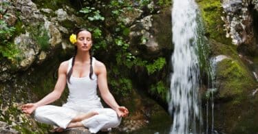 Imagem de uma mulher usando roupas brancas e uma rosa amarela no cabelo. Ela está fazendo a sua meditação e o seu mantra, sobre uma pedra, ao lado de uma pequena cachoeira.