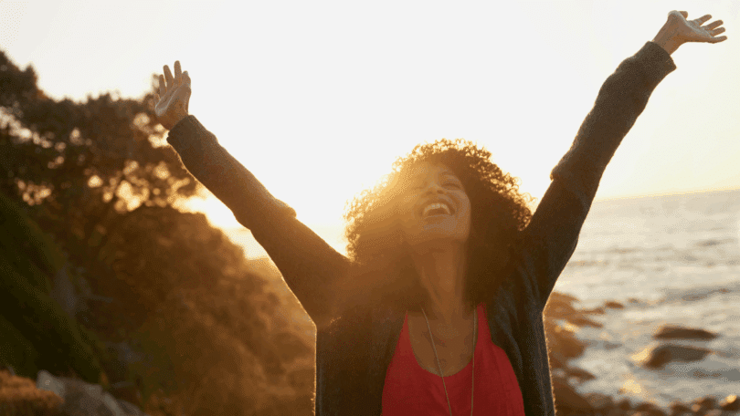 Mulher sorrindo com os braços erguidos ao pôr do sol à beira-mar.