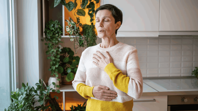 Mulher de olhos fechados respira profundamente com uma mão no peito e outra no abdômen, em uma cozinha com plantas ao fundo.