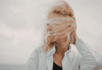 Mulher com cabelo ao vento cobrindo o rosto, usando camisa clara, em ambiente externo.