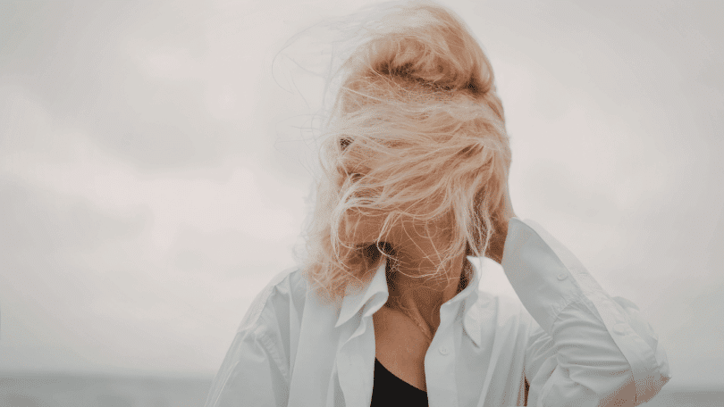 Mulher com cabelo ao vento cobrindo o rosto, usando camisa clara, em ambiente externo.