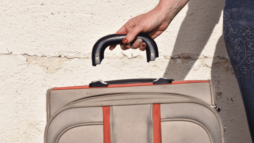 Mão segurando a alça de uma mala, destacada acima da própria bagagem.
